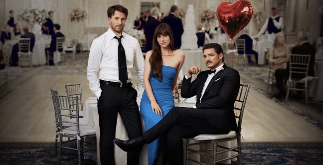 Three people in a room with a dance floor and baloons sat on wooden chairs. Two men wear suits, one with a dark blazer and a woman stands between them in a long blue dress.