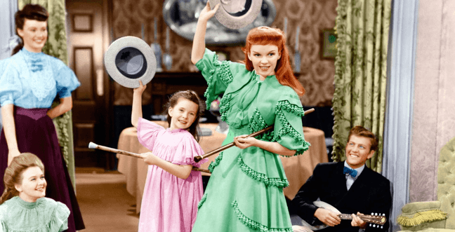 A group of people sit in a room with yellow curtains and pink wallpaper. A woman in a green dress with red hair holds a hat in the air with a stick under her arm next to a little girl in a pink dress