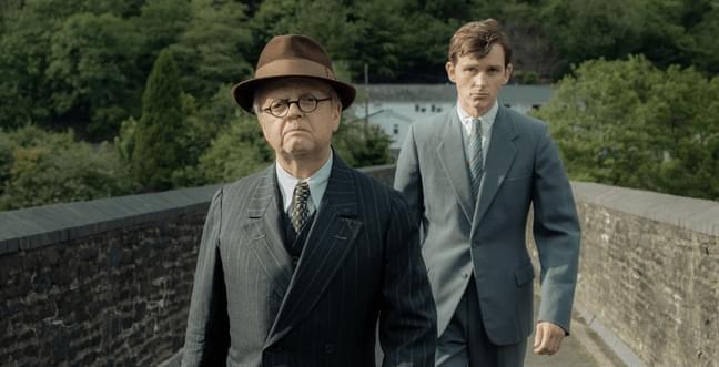 Two men in suits walk along a bridge, one wears a top hat and glasses and the other has short brown hair.