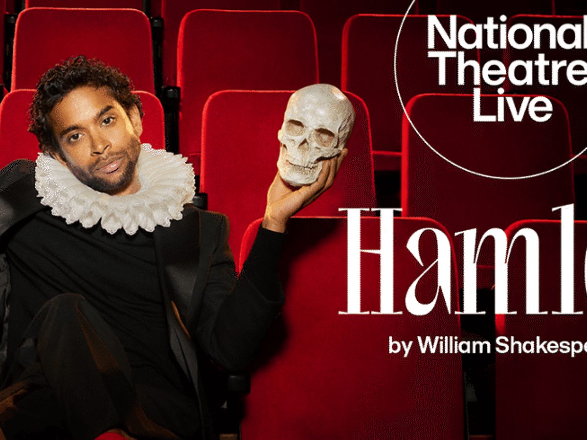 A man in black clothes and a white ruffle neck holding a skull while sitting on red theatre seats.