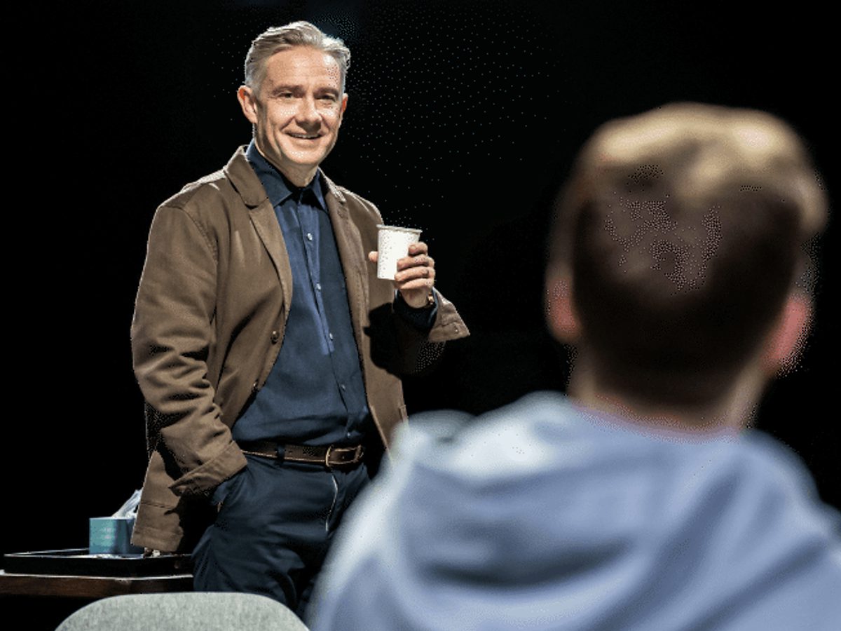 A man wearing a brown jacket and blue shirt smiles while holding a coffee cup, looking towards a man where we can only see the back of his head.