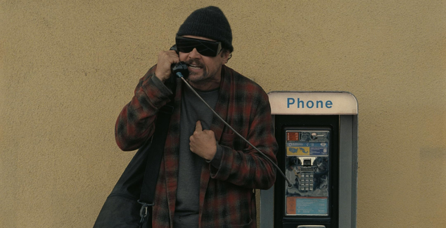 A man stands by a pay phone with dark glasses, a black hat and an open check shirt
