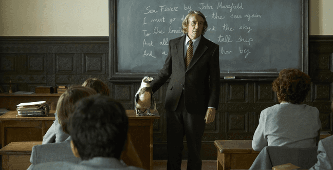 A man in front of a class room chalk board stands next to a penguin sat on a wooden desk