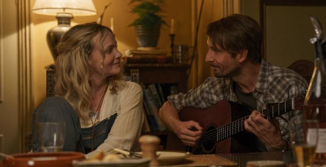 A man and a woman sat at a table looking into eachothers eyes. The man is holding a guitar