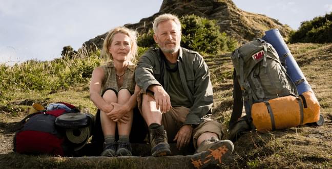 A middle aged man and woman sit on the grass of a hillside together surrounded by camping gear