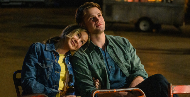 A blonde middle aged lady rests her head on the shoulder of a younger blonde man while sat outside at night