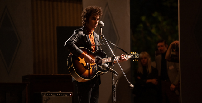 A man with a guitar in hand sits by a microphone with a leather jacket on