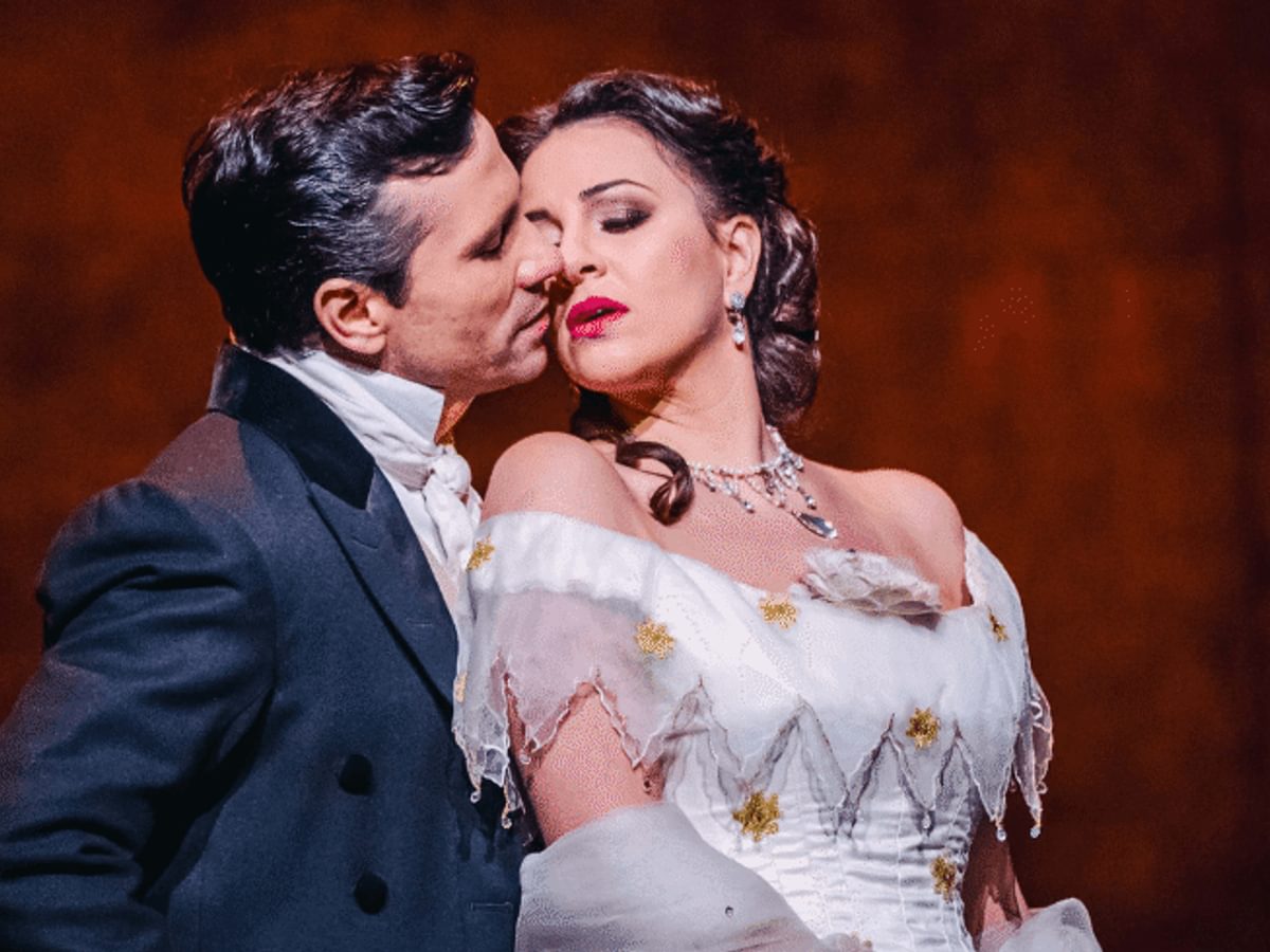 A man wearing a black suit embraces a woman from behind, who is wearing a white dress, standing in front of a red backdrop.