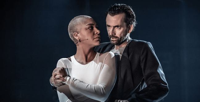 A black woman with a shaved head and a white mesh top is held in the arms of a white man with a brown beard and dark jumper on a dark stage, lit by a spotlight