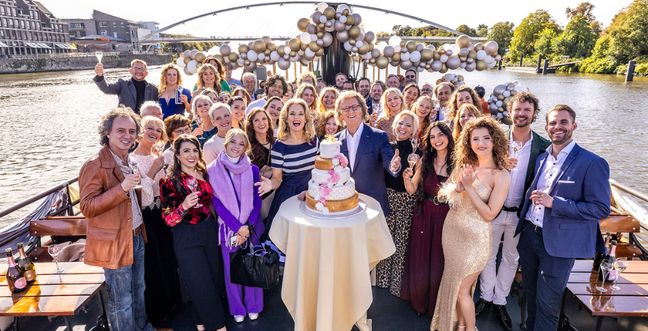 A group of people surrounded by gold, bronze and white balloons. In the foreground Andre Reiu stands with a tiered cake with the number 75 on top of it.
