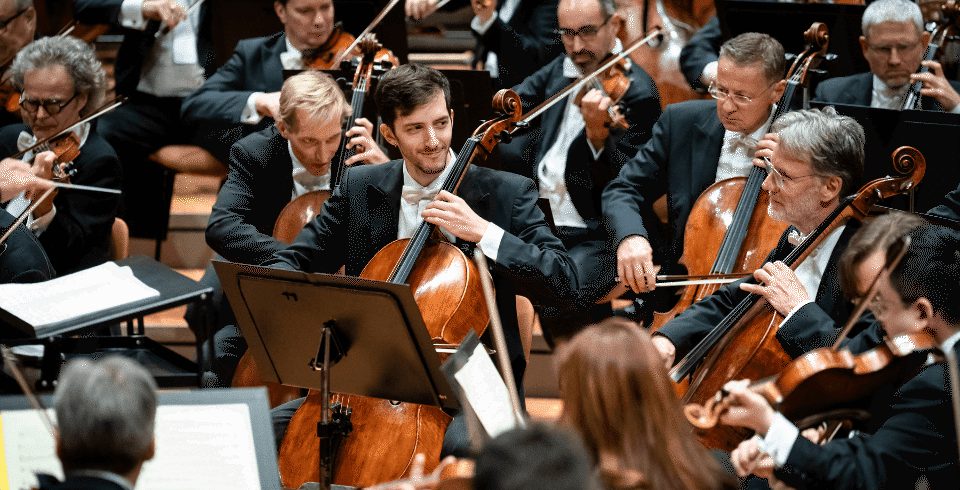A cellist standing among a seated string section within an orchestra. The musicians are wearing suits.