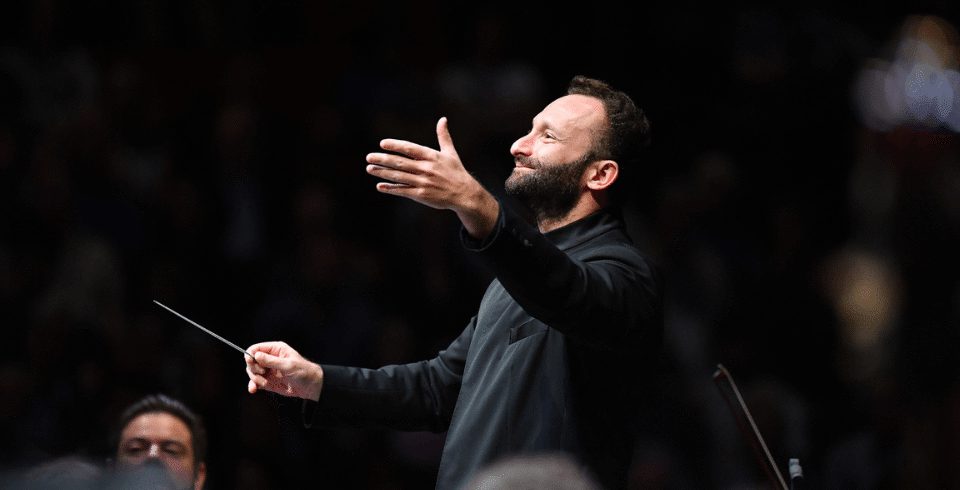 A conductor dressed in black, holding a baton, with his arms raised. He is smiling.