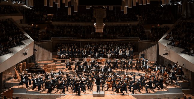 An orchestra within a philharmonic hall, holding their instruments with a conductor standing in front of them with their arms raised.
