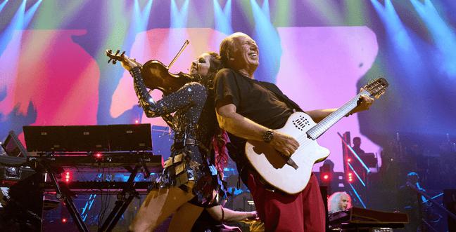 A man in red trousers smiles while holding a guitar stands back to back with a violinist wearing a sparkly silver dress. There are multi-coloured lights behind them