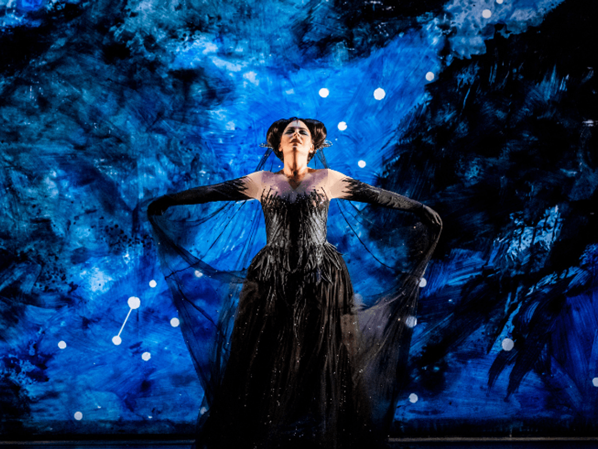 A woman in a black glittering mesh dress and gloves holds out the train of her dress behind her, lit by blue lights behind her