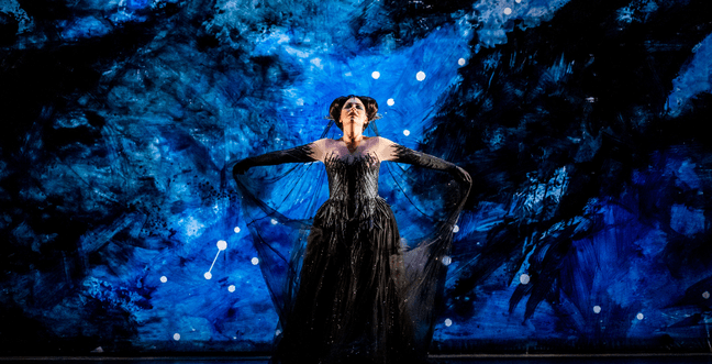 A woman in a black glittering mesh dress and gloves holds out the train of her dress behind her, lit by blue lights behind her