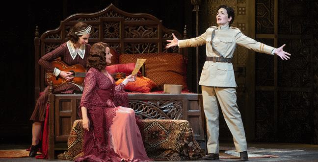 A woman sits at the end of a bed with a woman dressed as a maid in a red dress by her side and a woman in a uniform with their arms out stretched beside them