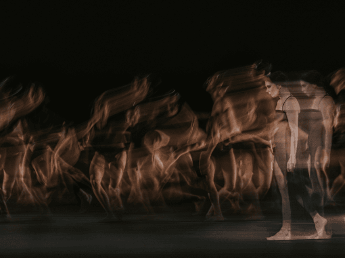 A black background with a group of dancers in motion, captured with a slow shutter speed to create a ghostly, blurred effect that emphasizes movement and fluidity.