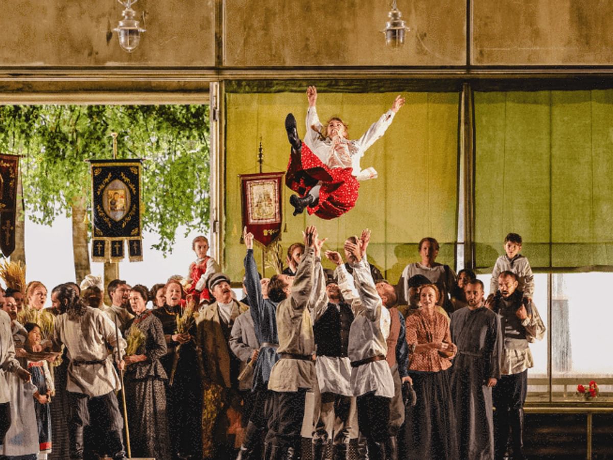 A girl in a red shirt and white top is thrown above a large crowd of people in a yellow room with large windows