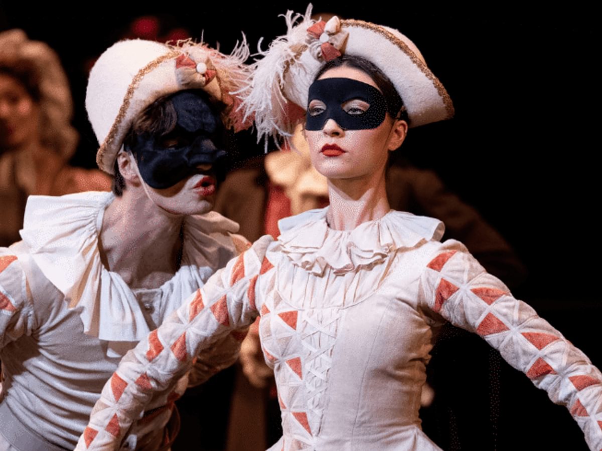 A man and a woman wearing black masks with white outfits and frilly collars stand on stage