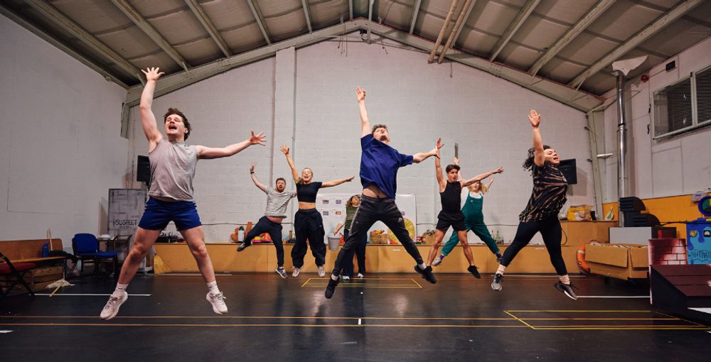 A group of people in a studio jump in the air with one hand pointing up and the other to the side