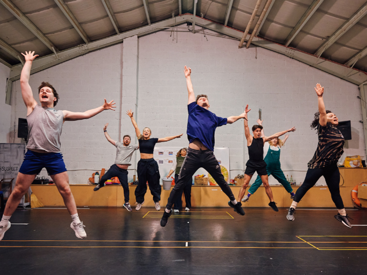 A group of people in a studio jump in the air with one hand pointing up and the other to the side