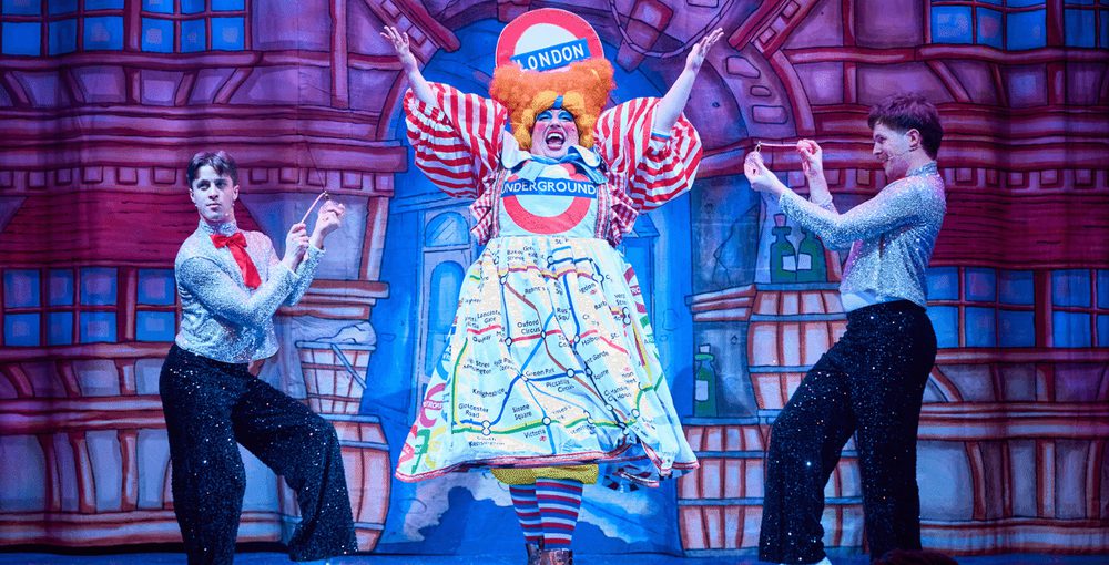 A pantomime dame dressed in a tube station print dress with a large London sign on their head stands next to two men in sparkly outfits holding perfume bottles