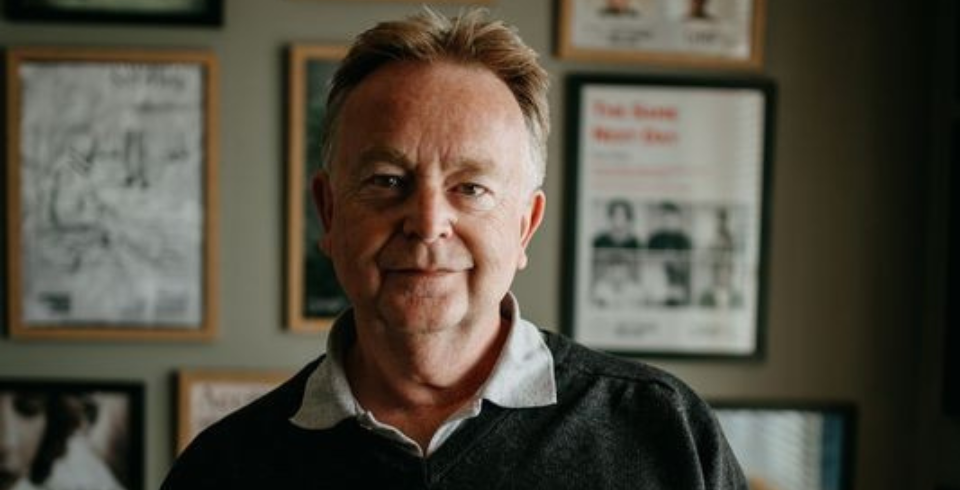 An older man stands in a study wearing a shirt and a V-neck knit jumper