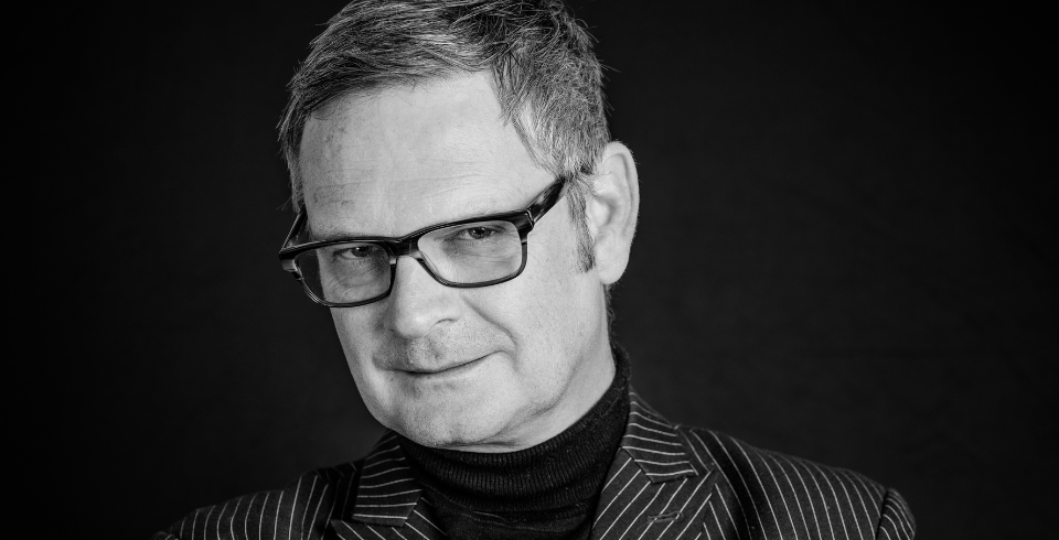 A man with rectangular glasses and a strong jaw, poses for the camera in a pinstriped suit