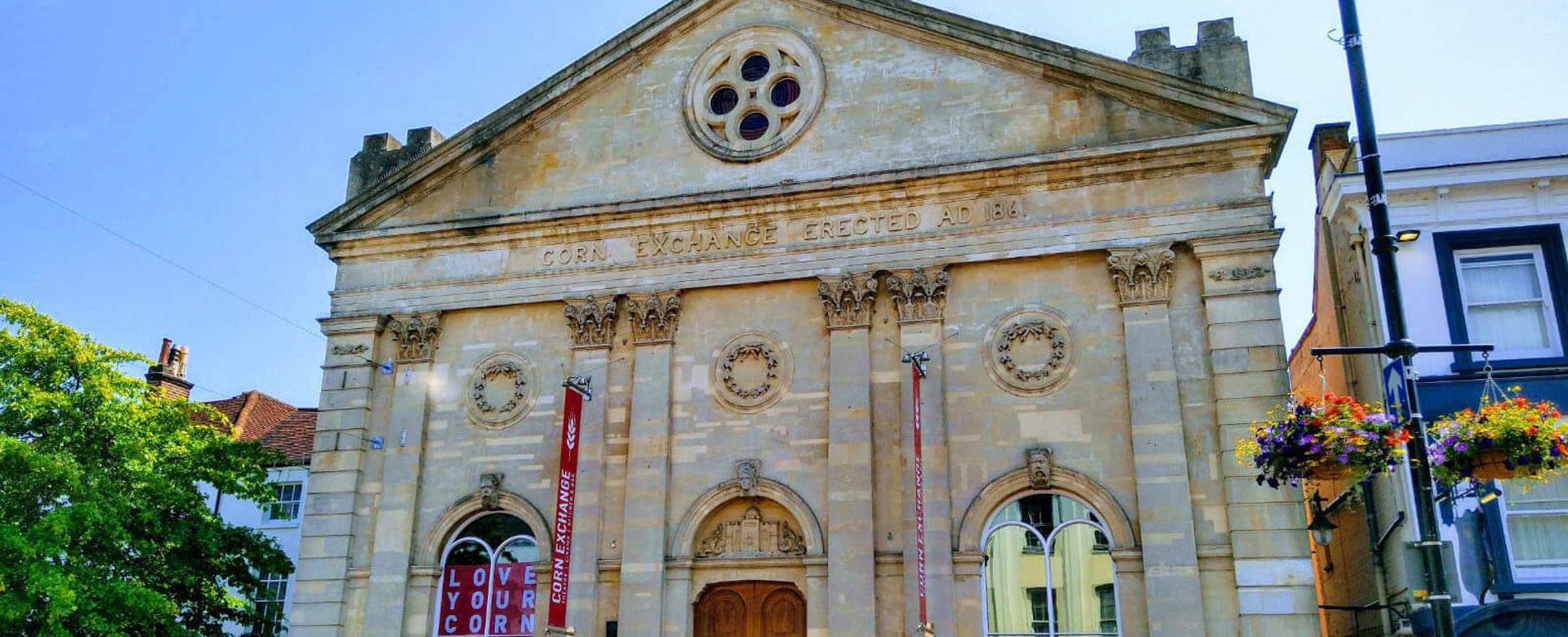 A stone building with a round window at the top and four pillars in the sunshine with blue sky.