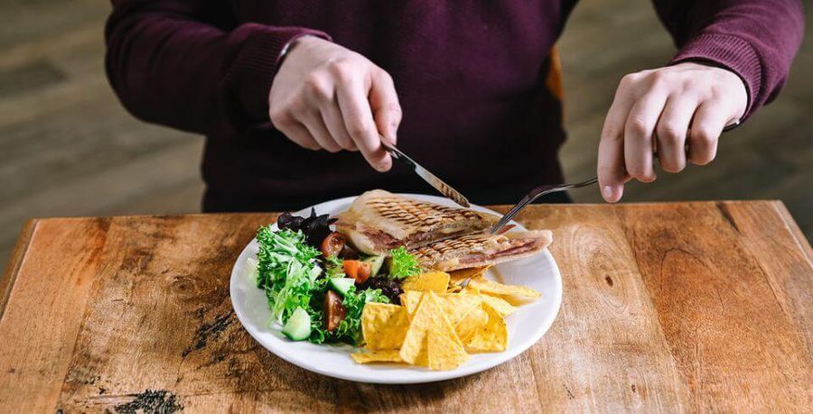 Hands cutting in to a ham panini with nacho chips on the side and a tomato salad set on a wooden table.