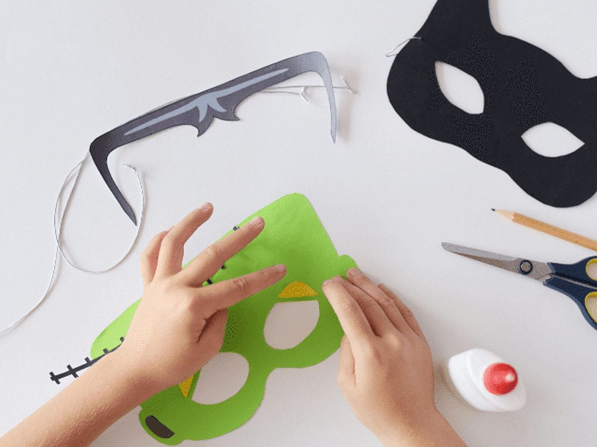 three handmade halloween masks on a white background. Two hands are editing one of them. a pair of scissors, pot of glue and a pencil are also in the shot.