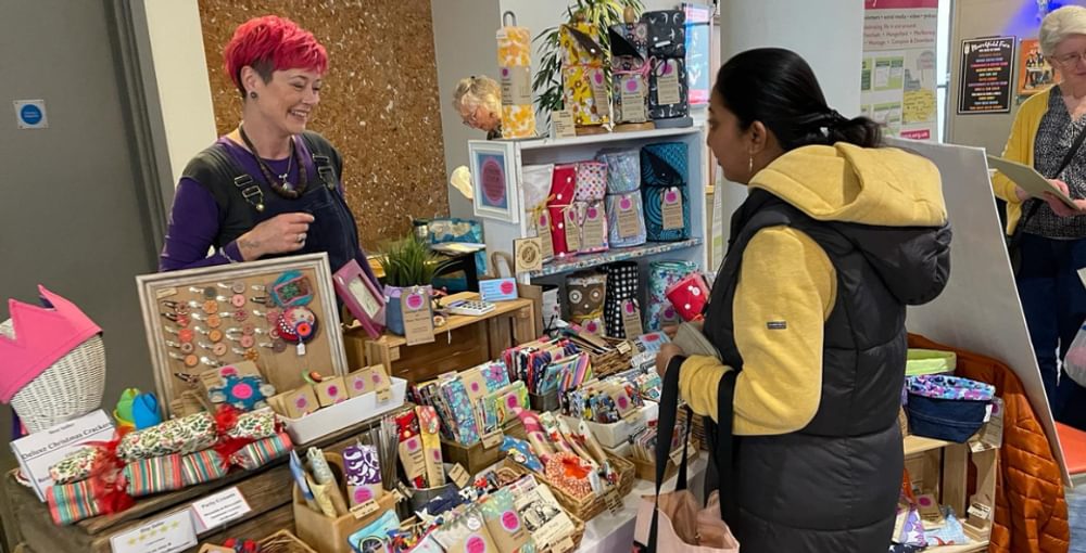 A woman with bright red short hair stands behind her stall of handmade items, talking to a customer