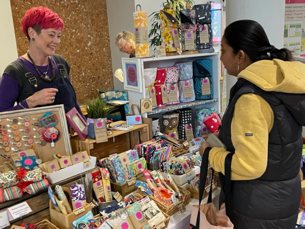 A woman with bright red short hair stands behind her stall of handmade items, talking to a customer