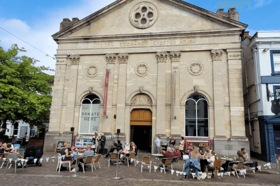 A large, old-fashioned building where people are sat outside on a sunny day.