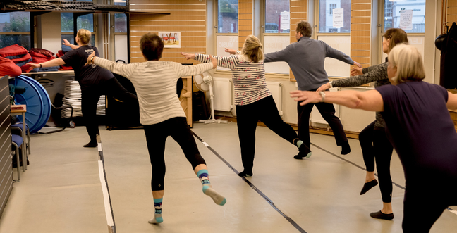 A group of people in a dance class stand on one leg with arms outstretched