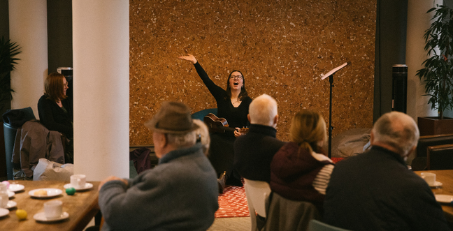 A woman sits in front of a group of people singing with one arm in the air