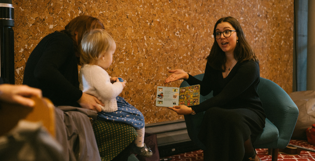 A woman holding a children's book sits in front of a child on a woman's lap