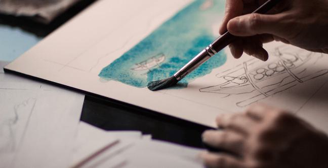 A hand holding a paintbrush, painting a watercolour turquoise line on a white piece of paper