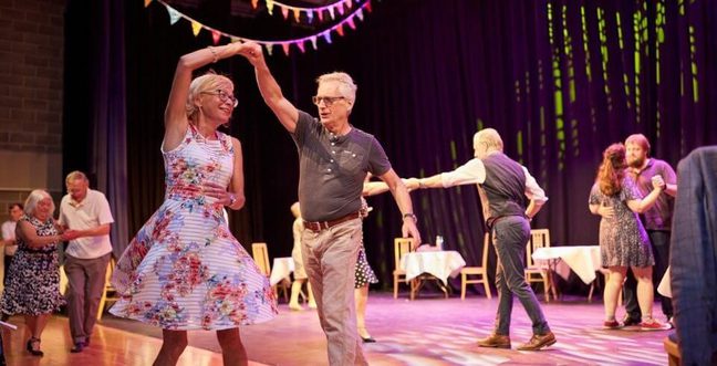 This is a photo of a room full of adults dancing. In the camera focus is a pair where the woman is mid-twirl in a swing dress.