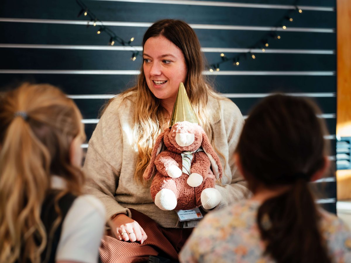 A woman holds a stuffed animal as she smiles and talks to children sat in front of her