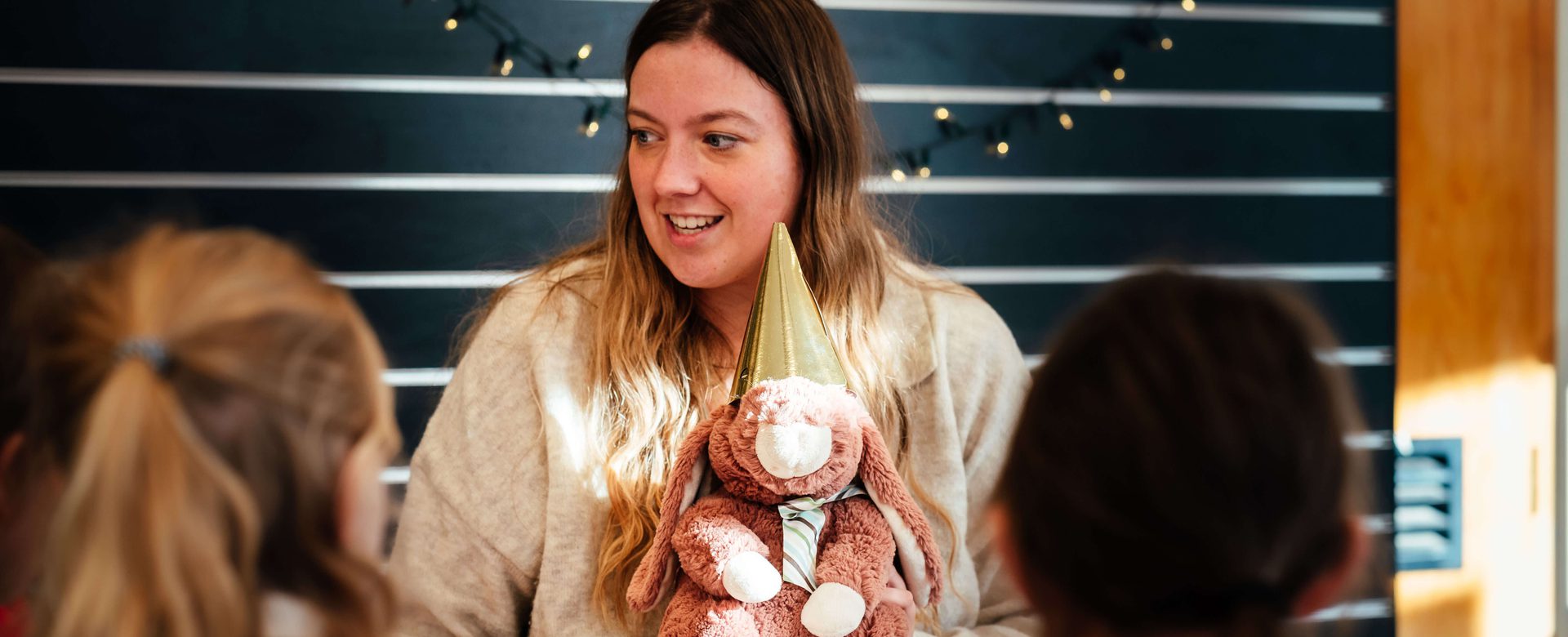 A woman holds a stuffed animal as she smiles and talks to children sat in front of her