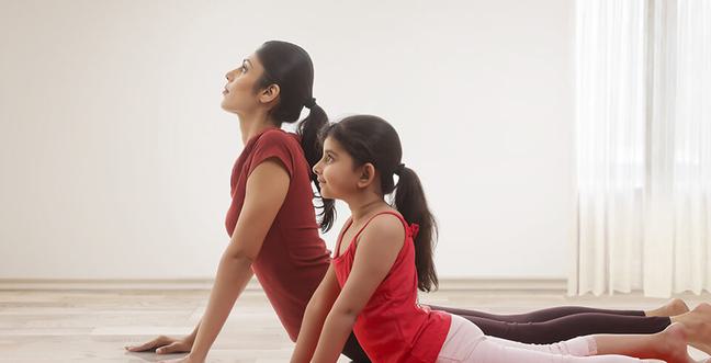 Two people, a parent and a child stretch in a yoga position wearing red clothing in a day-lit room