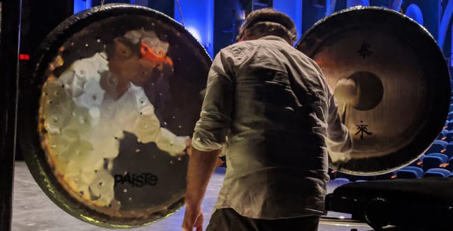 A man stands facing away from the camera, playing two large brass gongs.