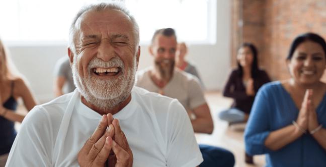 A man with his hands at heart centre smiles broadly while sat cross legged in a room of others doing the same