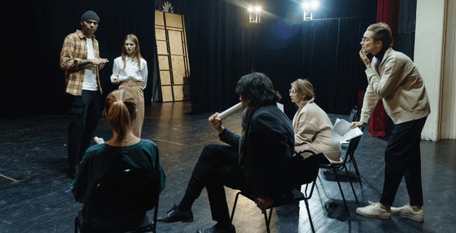 A group of actors stand around, some are sitting and some are stood and they are holding scripts