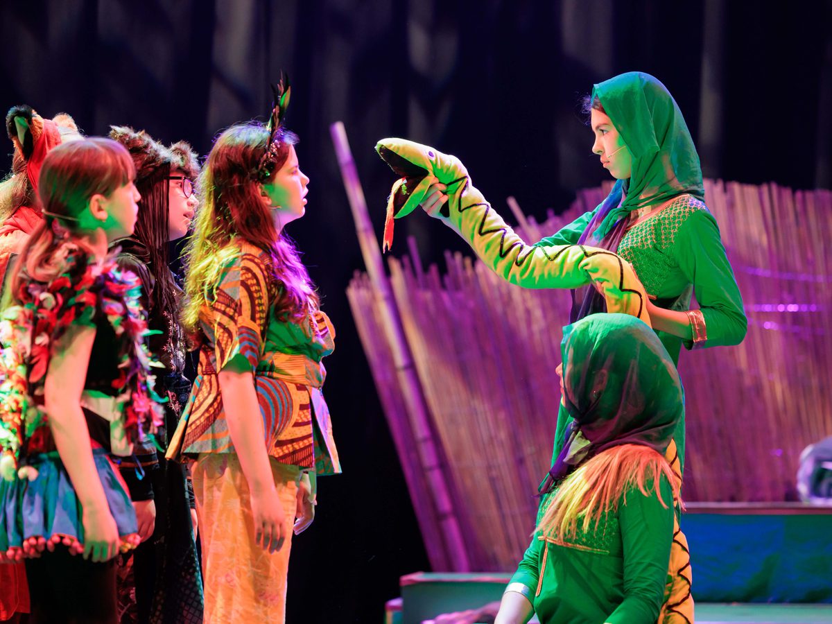 Two girls dressed in green holding a toy snake stand in front of a group of girls all dressed as various jungle animals