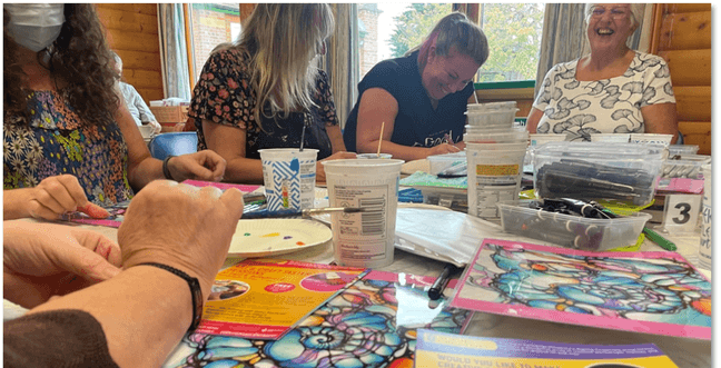 A group of women sit around a wooden table smiling and laughing while engaging in a craft activity