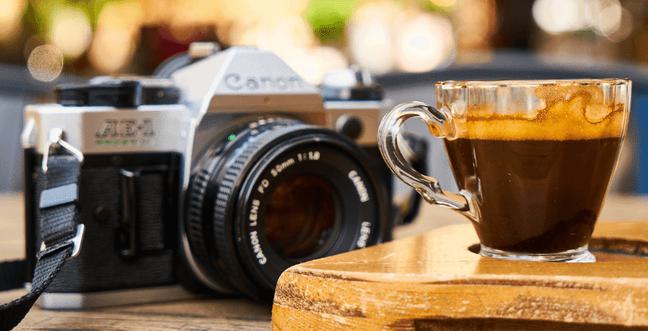 A film camera sits on a wooden table with a small coffee in front