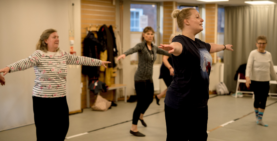 A group of people in a dance class hold their arms raised by their sides